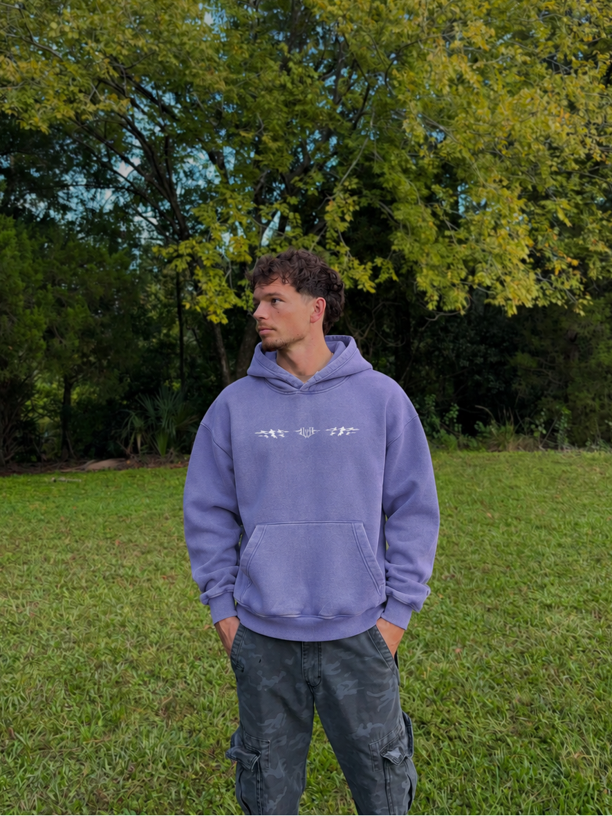 A man wearing a lavender-purple hoodie with a white graphic print on the chest and dark gray cargo pants, standing in a grassy park with green trees in the background.