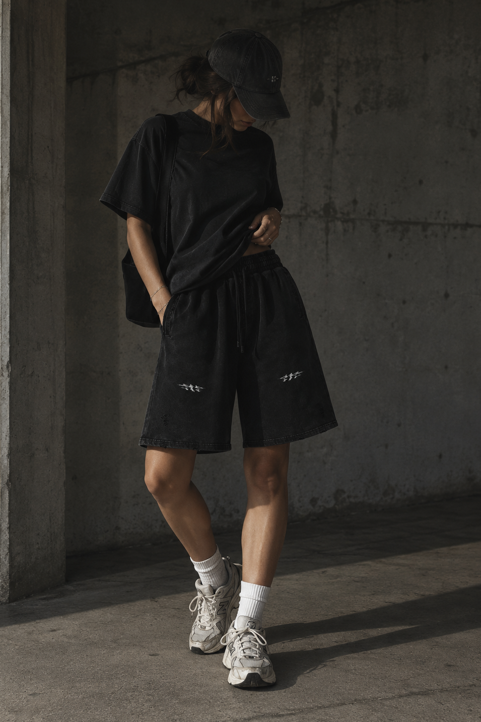 A person wearing a black oversized T-shirt, black shorts with small white logo prints, a black cap, white socks, and white sneakers, standing in a concrete industrial setting.